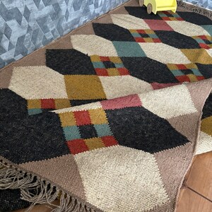 Pu&ograve; includere: Tappeto a fantasia geometrica con un mix di colori tra cui nero, beige, marrone, rosso e verde acqua. Il tappeto ha una frangia sul bordo e un motivo a rombi ripetuto. Un'auto decorativa gialla con una pianta &egrave; appoggiata sul tappeto.