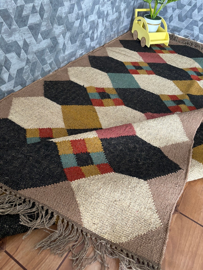 Pu&ograve; includere: Tappeto a fantasia geometrica con un mix di colori tra cui nero, beige, marrone, rosso e verde acqua. Il tappeto ha una frangia sul bordo e un motivo a rombi ripetuto. Un'auto decorativa gialla con una pianta &egrave; appoggiata sul tappeto.
