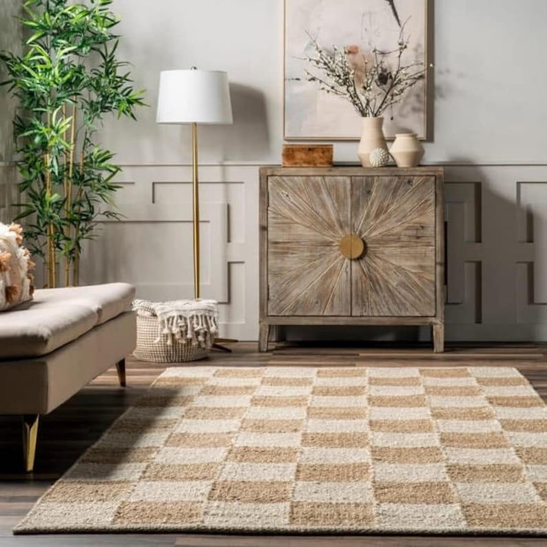 May include: A beige and white checkered jute rug with a natural woven texture. The rug is in a living room setting with a sofa, a floor lamp, and a wooden cabinet.