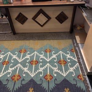 May include: A blue, white, red, and yellow patterned rug with fringe. The rug has a geometric design and is laid out on a tiled floor.