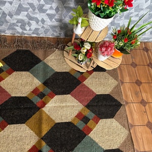 Pu&ograve; includere: Un tappeto a motivi geometrici con un design esagonale ripetuto in nero, beige, rosso e verde acqua. Il tappeto &egrave; frangiato su un lato. Sono visibili anche fiori artificiali in vasi bianchi e un portapiante a pi&ugrave; livelli in legno.