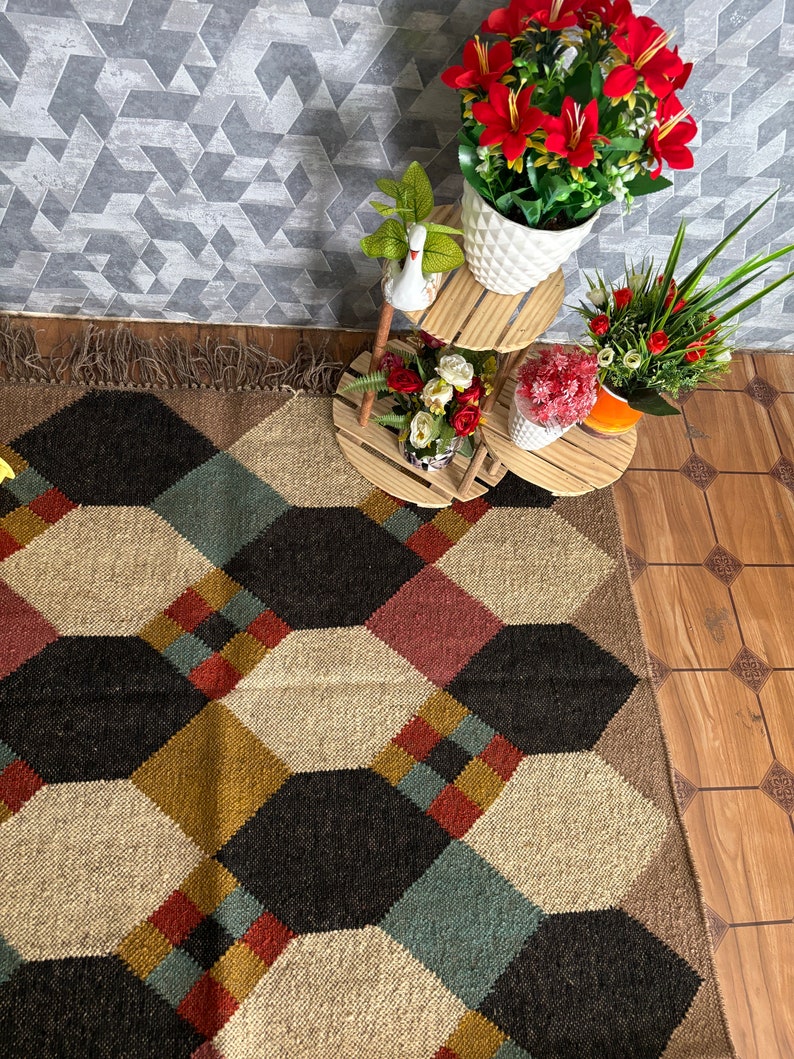 Pu&ograve; includere: Un tappeto a motivi geometrici con un design esagonale ripetuto in nero, beige, rosso e verde acqua. Il tappeto &egrave; frangiato su un lato. Sono visibili anche fiori artificiali in vasi bianchi e un portapiante a pi&ugrave; livelli in legno.