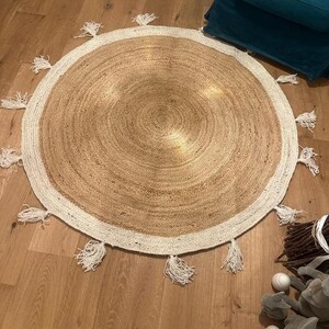 Puede incluir: Una alfombra redonda de yute con un borde blanco y flecos. La alfombra tiene un tono natural y terroso y es perfecta para agregar un toque de estilo bohemio a cualquier habitaci&oacute;n.