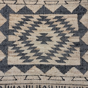 May include: Black and beige woven rug with a geometric pattern. The rug has a central square with a black and white geometric design. The border of the rug is a black and beige geometric pattern.