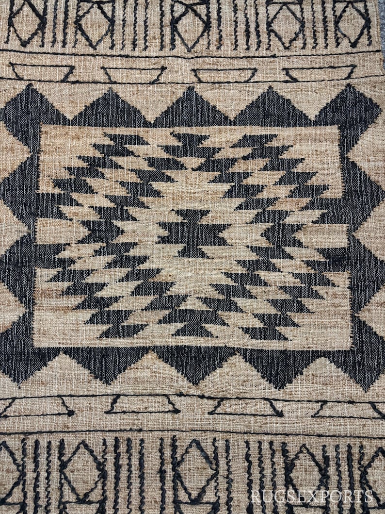 May include: Black and beige woven rug with a geometric pattern. The rug has a central square with a black and white geometric design. The border of the rug is a black and beige geometric pattern.