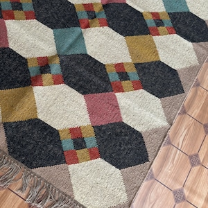 Pu&ograve; includere: Tappeto a motivi geometrici con un design a cubi ripetuti in nero, crema, giallo senape, verde acqua e rosa. Il tappeto ha un bordo con frange ed &egrave; posto su un pavimento in legno.