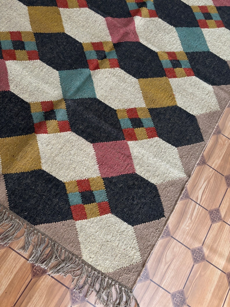Pu&ograve; includere: Tappeto a motivi geometrici con un design a cubi ripetuti in nero, crema, giallo senape, verde acqua e rosa. Il tappeto ha un bordo con frange ed &egrave; posto su un pavimento in legno.