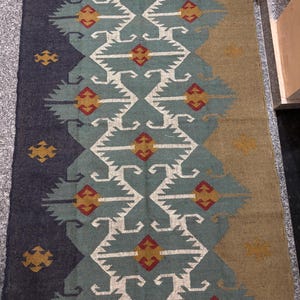 May include: A blue, green, and brown patterned rug with a geometric design. The rug has a fringe border.