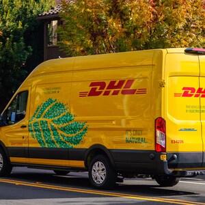 Pu&ograve; includere: Un furgone DHL giallo brillante con il logo DHL rosso. Il testo "It's Only Natural" e un motivo a foglia verde adornano il lato. Il veicolo &egrave; elettrico. Lo sfondo mostra alberi e case.