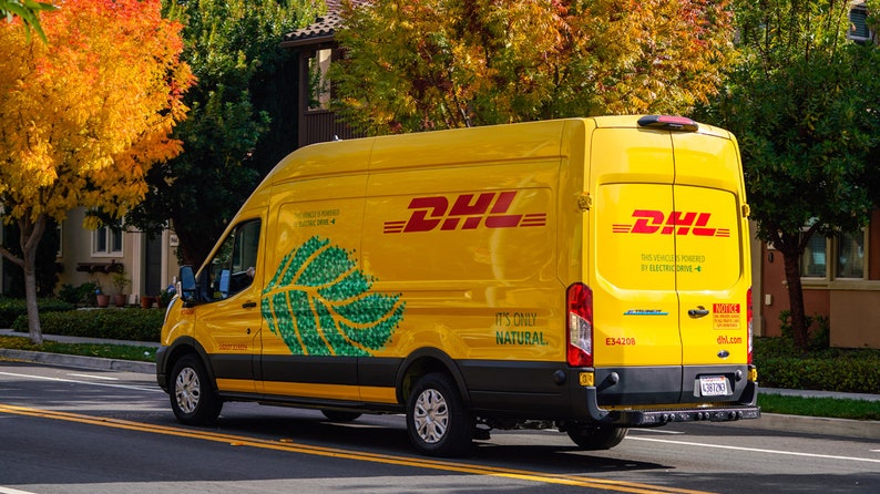 Pu&ograve; includere: Un furgone DHL giallo brillante con il logo DHL rosso. Il testo "It's Only Natural" e un motivo a foglia verde adornano il lato. Il veicolo &egrave; elettrico. Lo sfondo mostra alberi e case.