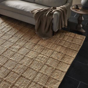 May include: A natural fiber rug with a woven, checkered pattern in a light brown color. The rug is made of jute and has a natural, earthy look.