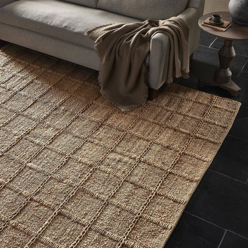 May include: A natural fiber rug with a woven, checkered pattern in a light brown color. The rug is made of jute and has a natural, earthy look.
