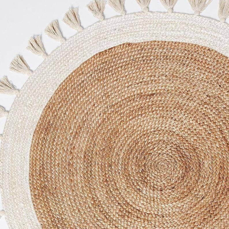 Puede incluir: Una alfombra redonda de yute con un borde blanco y flecos. La alfombra tiene un tono natural y terroso y es perfecta para agregar un toque de estilo bohemio a cualquier habitaci&oacute;n.