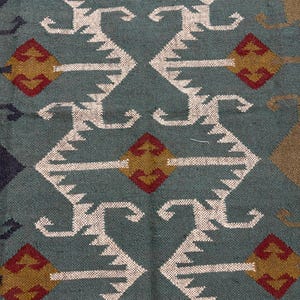 May include: A teal blue rug with a geometric pattern in white, red, and gold. The pattern features repeating diamond shapes and stylized curves.