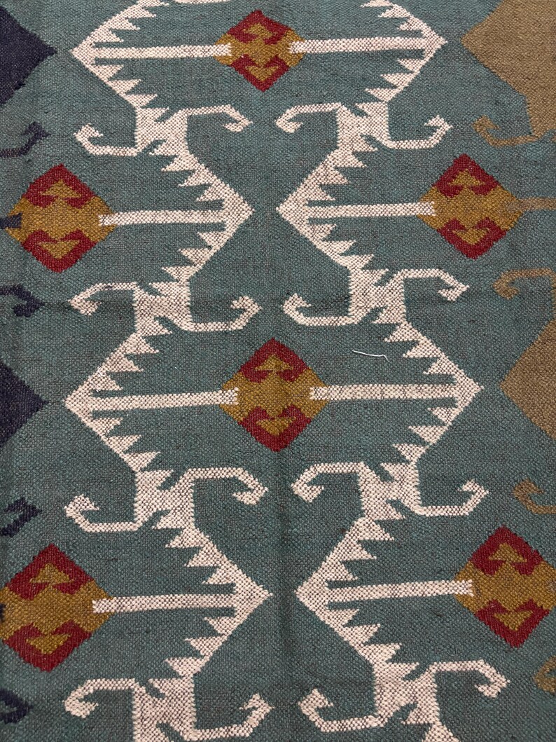 May include: A teal blue rug with a geometric pattern in white, red, and gold. The pattern features repeating diamond shapes and stylized curves.