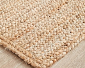 May include: A rectangular area rug crafted from natural tan jute fibers, featuring a dense, uniform braided weave pattern. The rug displays a consistent light brown, earthy tone throughout, with visible fiber texture and a sturdy, thick border.
