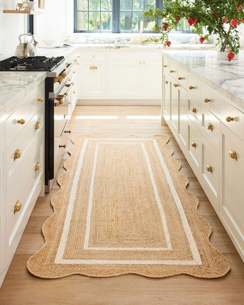 May include: A rectangular jute rug with a scalloped edge and a white border. The rug is placed in a kitchen with white cabinets, a black stove, and light wood flooring. The rug adds a natural, textured element to the space.