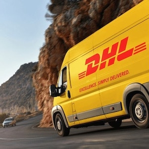 May include: A yellow DHL delivery van with red lettering and the text "EXCELLENCE. SIMPLY DELIVERED." is driving on a winding road. The van is on a road next to a rocky cliffside.