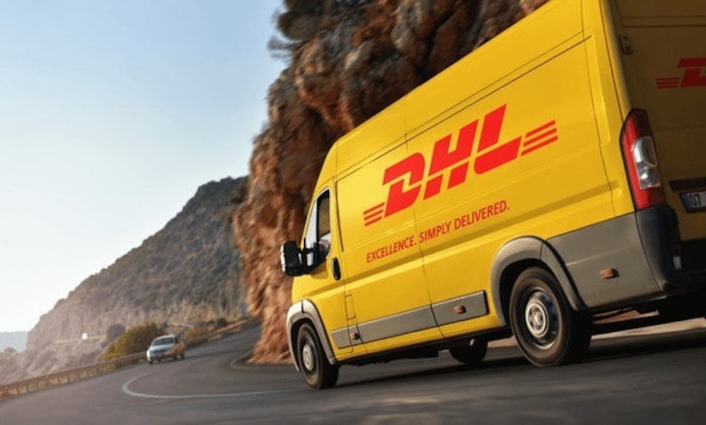 May include: A yellow DHL delivery van with red lettering and the text "EXCELLENCE. SIMPLY DELIVERED." is driving on a winding road. The van is on a road next to a rocky cliffside.