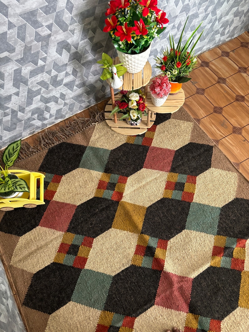 Pu&ograve; includere: Tappeto a motivi geometrici con un motivo esagonale ripetuto in nero, beige, rosso e verde acqua. Il tappeto ha un bordo sfrangiato ed &egrave; posto su un pavimento in legno. Vasi di fiori decorativi su supporti in legno.