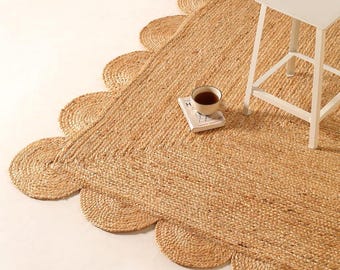 May include: A rectangular natural tan jute rug featuring a decorative scalloped edge design. A white four-legged stool stands on the rug, and a small white book with a dark beverage in a ceramic mug is placed on the textured woven surface.