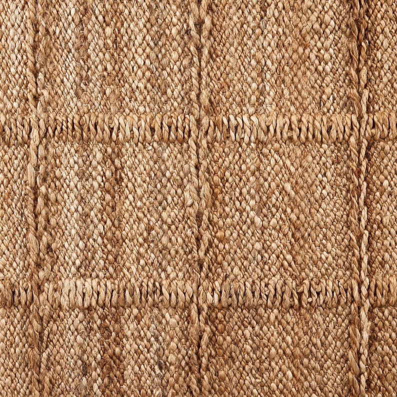 May include: Close-up of a woven jute rug with a natural brown color and a textured, slightly rough surface. The rug has a grid pattern created by thicker woven lines.