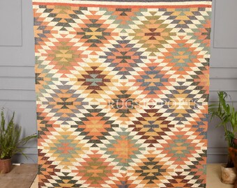 May include: A rectangular woven rug featuring a repeating diamond pattern in shades of orange, green, blue, and cream. The rug has dark blue borders at the top and bottom with a row of small cross motifs and frayed fringe edges at both ends.