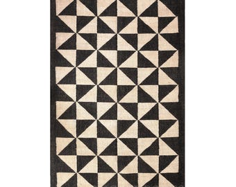 May include: A rectangular area rug featuring a repeating geometric pattern of black and off-white triangles arranged in a grid. The rug is finished with short, light-colored fringe tassels along the top and bottom edges, creating a textured border.