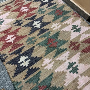 Puede incluir: Una alfombra rectangular con un patr&oacute;n geom&eacute;trico en tonos beige, rojo, verde, azul marino y rosa. La alfombra presenta un dise&ntilde;o repetitivo de formas estilizadas en forma de cruz. El texto "RUGSEXPORTS" es visible en la esquina inferior derecha.
