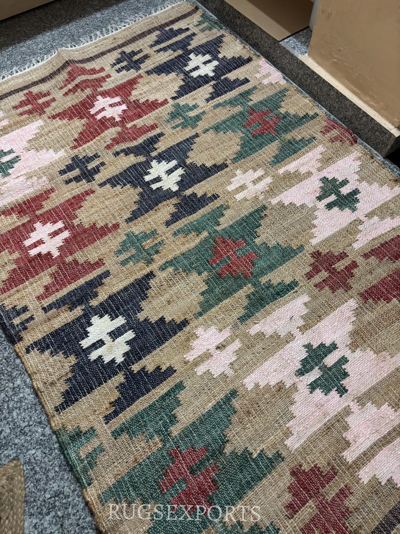 Puede incluir: Una alfombra rectangular con un patr&oacute;n geom&eacute;trico en tonos beige, rojo, verde, azul marino y rosa. La alfombra presenta un dise&ntilde;o repetitivo de formas estilizadas en forma de cruz. El texto "RUGSEXPORTS" es visible en la esquina inferior derecha.