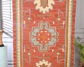 May include: A rectangular woven rug featuring a terracotta red background with a central geometric medallion in light blue and cream. The border displays a zigzag pattern in cream and blue, with additional symmetrical motifs in olive green.