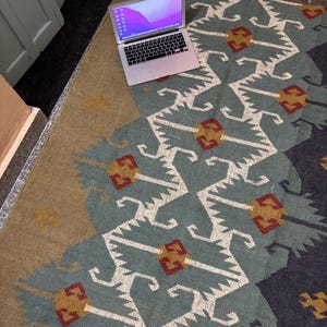 May include: A laptop computer with a light purple and blue gradient background on the screen is sitting on a rug with a geometric pattern in shades of blue, white, red, and gold.
