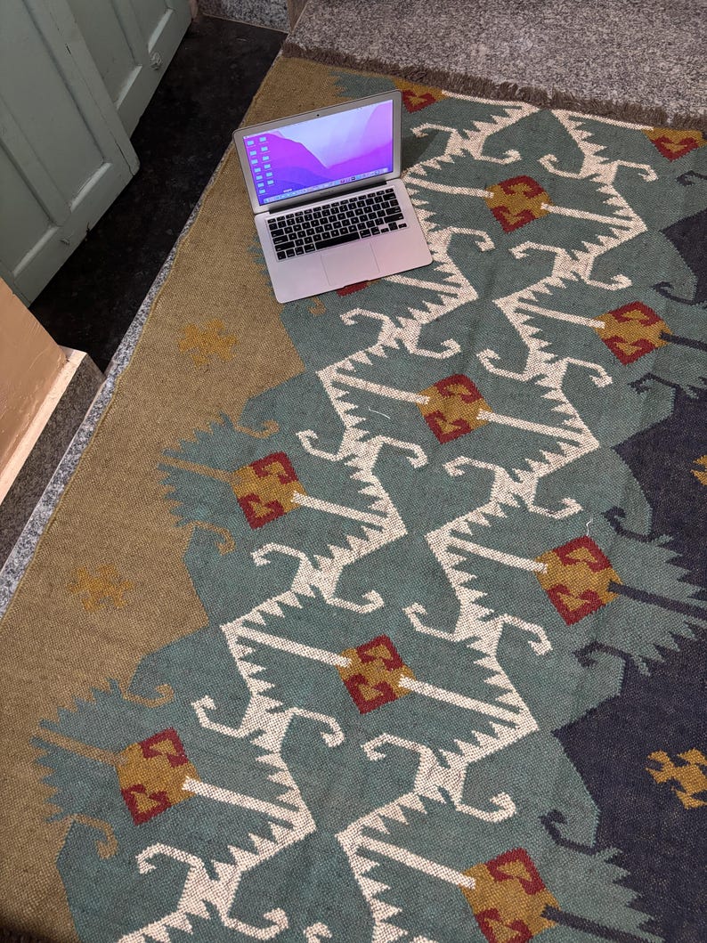 May include: A laptop computer with a light purple and blue gradient background on the screen is sitting on a rug with a geometric pattern in shades of blue, white, red, and gold.