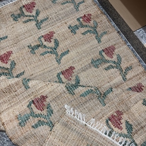 Puede incluir: Alfombra de yute beige con un patr&oacute;n repetido de dise&ntilde;os florales rojos y tallos verdes. La alfombra tiene un borde con flecos. El texto "RUGSEXPORTS" es visible en la imagen.
