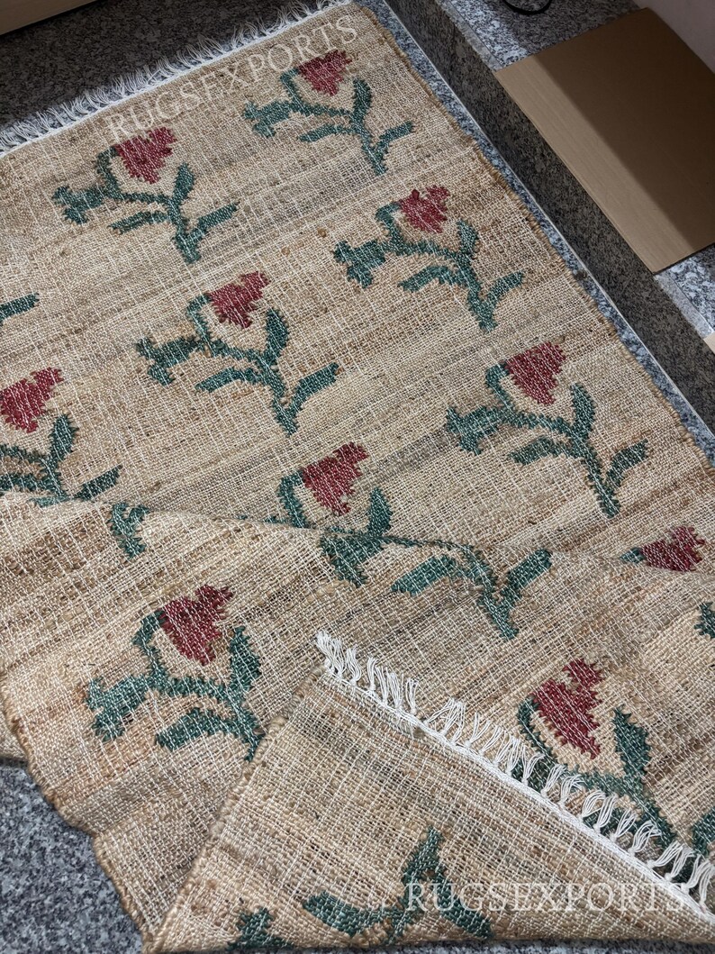 Puede incluir: Alfombra de yute beige con un patr&oacute;n repetido de dise&ntilde;os florales rojos y tallos verdes. La alfombra tiene un borde con flecos. El texto "RUGSEXPORTS" es visible en la imagen.
