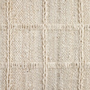 May include: Close-up of a cream-colored woven rug with a textured surface. The rug has a subtle pattern of raised squares.