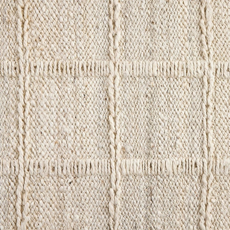May include: Close-up of a cream-colored woven rug with a textured surface. The rug has a subtle pattern of raised squares.