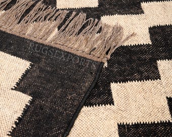 May include: A woven rug featuring a repeating black and cream geometric checkerboard pattern. The rug has a textured surface and includes a fringe edge made of light brown fibers. The text RUGSEXPORTS is visible on the surface of the rug.