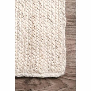 Puede incluir: Primer plano de una alfombra tejida blanca con una superficie texturizada. La alfombra está hecha de fibras naturales y tiene un diseño simple y elegante.