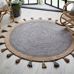 Puede incluir: Una alfombra redonda de yute gris y beige con un borde de flecos. La alfombra tiene una textura tejida y un aspecto de fibra natural.