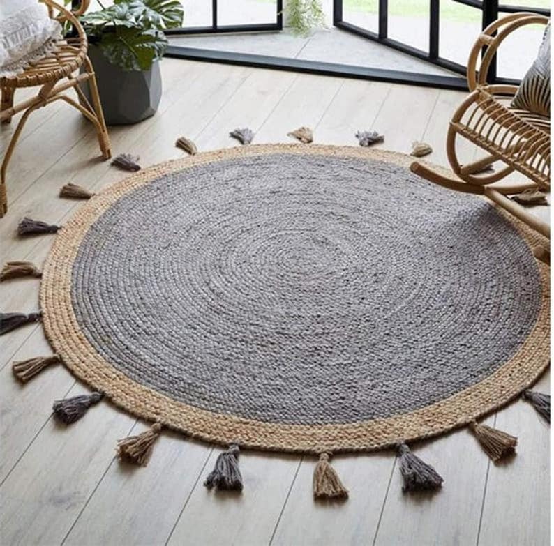 Puede incluir: Una alfombra redonda de yute gris y beige con un borde de flecos. La alfombra tiene una textura tejida y un aspecto de fibra natural.
