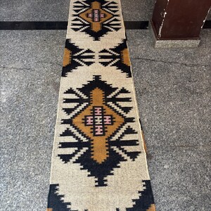 May include: A rectangular beige runner rug with a geometric pattern in black, brown, and pink. The rug has a repeating diamond and zigzag design. The rug is made of woven material and is placed on a gray surface.