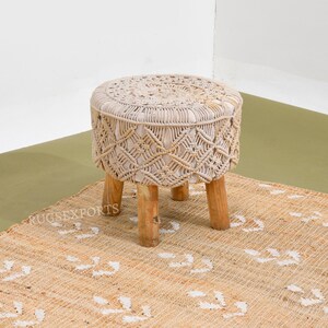 May include: A white macrame stool with a round top and four wooden legs. The stool is sitting on a natural fiber rug with a white pattern.