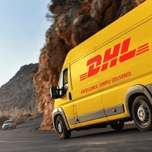 K&ouml;nnte beinhalten: Ein gelber Lieferwagen mit dem DHL-Logo und dem Slogan "Excellence. Simply delivered." f&auml;hrt auf einer kurvigen Stra&szlig;e.