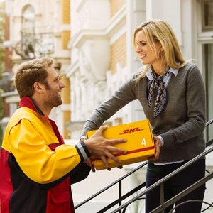 May include: A DHL delivery person in a yellow and red uniform hands a yellow box with the DHL logo to a woman. The woman is standing on a porch, and the delivery is taking place in front of a brick building.