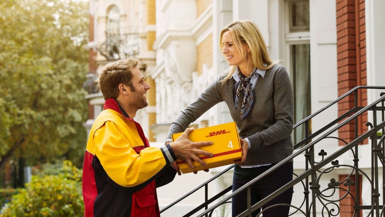 May include: A DHL delivery person in a yellow and red uniform hands a yellow box with the DHL logo to a woman. The woman is standing on a porch, and the delivery is taking place in front of a brick building.