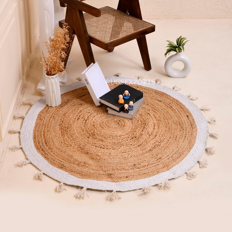 Puede incluir: Una alfombra redonda de yute con un borde blanco y flecos. La alfombra est&aacute; sobre un suelo de madera clara y hay peque&ntilde;as figuritas decorativas encima.