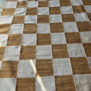 May include: A checkered rug with alternating squares of brown and white. The rug is made of a woven material and has a slightly textured surface.