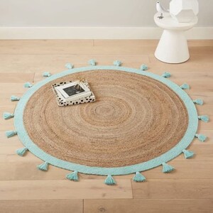 Puede incluir: Una alfombra redonda de yute con un borde azul claro y borlas. La alfombra tiene una textura natural y tejida.