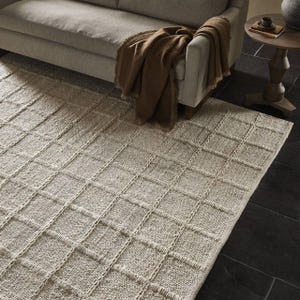 May include: A cream-colored woven rug with a grid pattern. The rug is placed under a light brown sofa with a brown throw blanket.